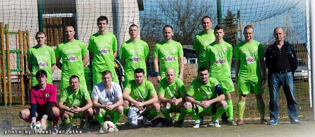 Gruppe von Männern in grünen T-Shirts, die auf einem Fußballfeld stehen oder knien, mit einem Ball davor, einem Torpfosten rechts daneben und diversen Hintergrundelementen wie Bäumen, Gebäuden und Fahrzeugen.