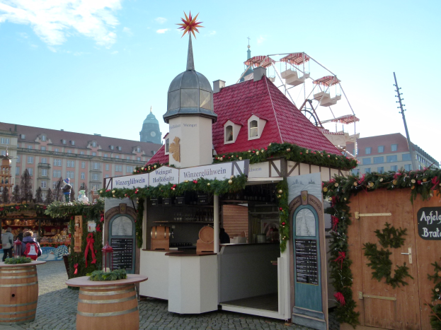 Kleines Gebäude mit rotem Dach und Riesenrad davor, umgeben von Festdekorationen und Menschen, vor einem Hintergrund aus Gebäuden, Bäumen und einem bewölkten Himmel während des Oktoberfestes in München, Deutschland.