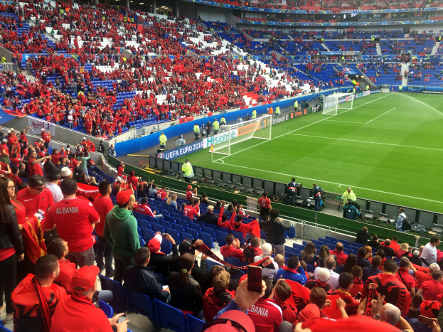 Ein volles Stadion mit Zuschauern, einige sitzen, einige stehen, mit Bannern, einem Torpfosten und Deckenleuchten, die gespannt auf den Beginn eines Fussballspiels warten.