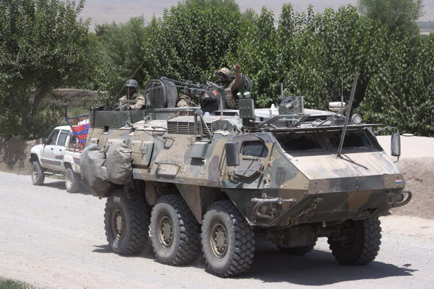Ein gepanzertes Personentransportfahrzeug und ein Lastwagen fahren eine Schotterstraße entlang, mit bewaffneten Personen in Helmen im Hintergrund.