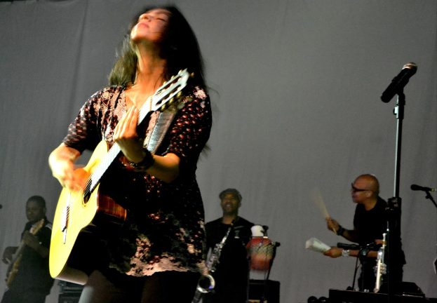 Eine Frau, die auf der Bühne eine Gitarre spielt, mit Musikern und verschiedenen Musikinstrumenten im Hintergrund und einem Mikrofon rechts.