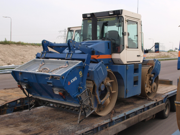 Ein Walzenfahrzeug wird auf einen Tieflader geladen, neben der Straße mit Laternenmasten, Bäumen und einem klaren blauen Himmel im Hintergrund.
