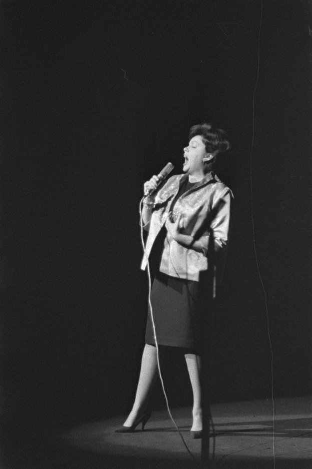 Eine Frau in einem schwarzen und weißen Outfit aus den 1950er Jahren singt fröhlich auf der Bühne unter einem Scheinwerfer und hält ein Mikrofon.