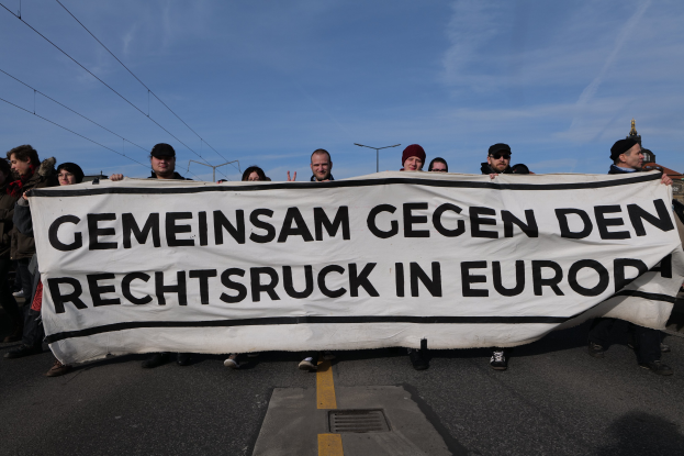 Eine Gruppe von Menschen auf einer Straße mit einem Banner, auf dem 'Gemeinsam gegen den Rechtsruck in Europa' steht, vor Hintergrund von Straßenlaternen, Strommasten, Stromkabeln, Gebäuden und einem bewölkten Himmel