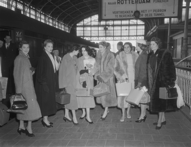 Schwarzes und weißes Bild einer Gruppe von Frauen, die zusammen auf einem Bahnhof stehen, jede hält eine Tasche in der Hand, mit einem Zug auf der linken Seite und Geländern und einer Tafel auf der rechten Seite.