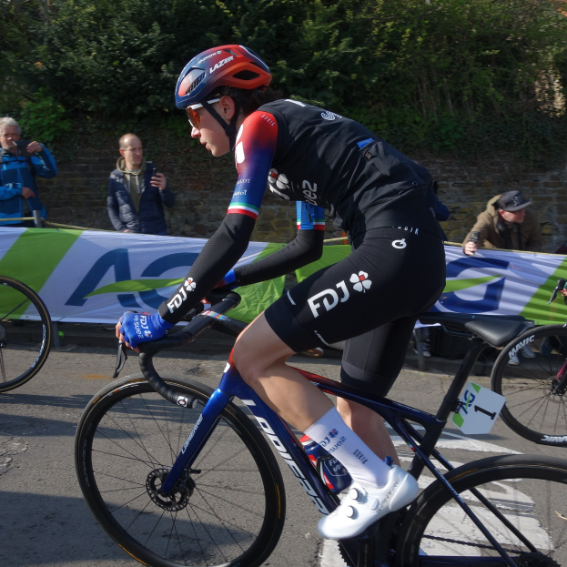 Professionelle Radrennfahrerin Katie Archibald trägt einen Helm, Schutzbrille, Handschuhe und Schuhe und fährt ein Fahrrad eine Straße entlang neben einer Menge, mit einem Banner, einer Wand, Bäumen und einem Gebäude im Hintergrund.