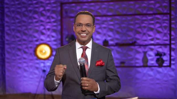 Stephen Colbert, der in Anzug, Krawatte und Brille auf einer Bühne steht und ein Mikrofon hält, mit einer Wand, einem Ständer, einer Lampe und verschiedenen Gegenständen im Hintergrund.