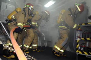 Feuerwehrleute in Schutzausrüstung und Helmen arbeiten in der Nähe eines Feuerwehrautos, mit Ausrüstungsgestellen und Hintergrundinfrastruktur.