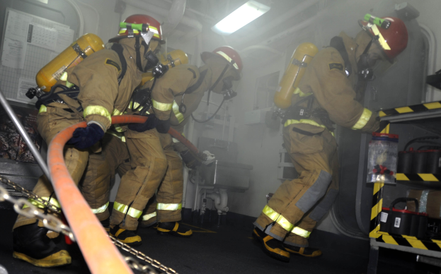 Feuerwehrleute in Schutzausrüstung und Helmen arbeiten in der Nähe eines Feuerwehrautos, mit Ausrüstungsgestellen und Hintergrundinfrastruktur.