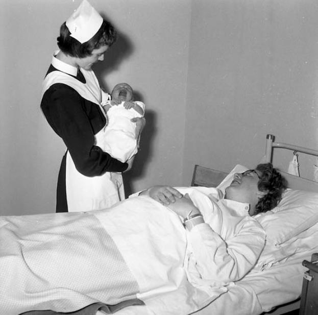 Eine Frau liegt in einem Krankenhausbett, das mit einer Decke und Kissen bedeckt ist, während eine Schwester neben ihr steht und ein Baby hält, mit einem Tisch mit verschiedenen Gegenständen rechts neben dem Bett, alles in Schwarz-Weiß dargestellt.