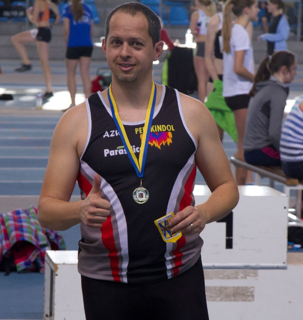 Mann in schwarzem Trikot und Medaille posiert, während im Hintergrund einige Menschen trainieren.