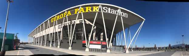Großes Stadiongebäude mit Text, umgeben von Pfählen, Lampen, Bäumen und einem Zaun, mit Himmel im Hintergrund.