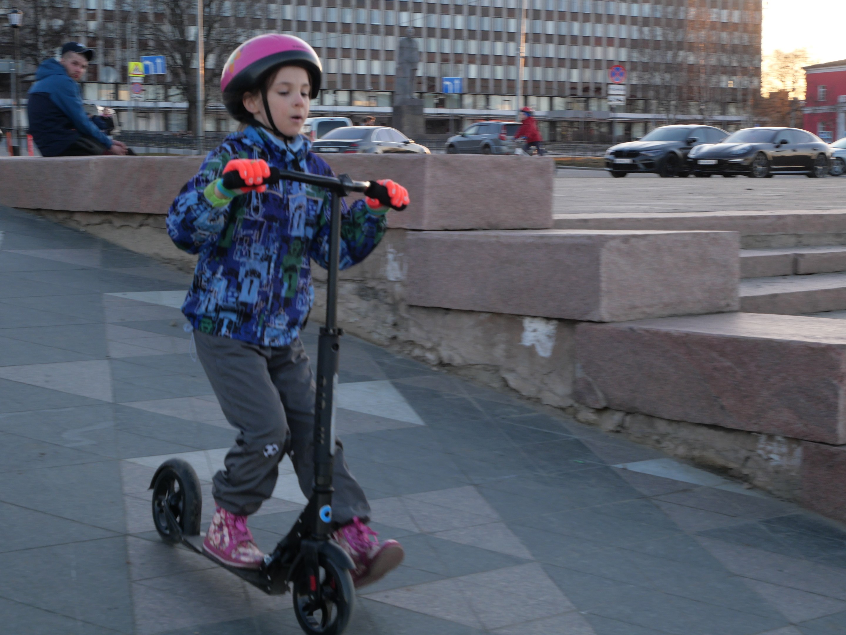 Ein junger Junge in einem Helm und Handschuhen fährt mit einem Roller eine Treppe auf einem Gehweg herunter, mit Fahrzeugen, Menschen, Bäumen, Polen, Brettern, Gebäuden und einem klaren blauen Himmel im Hintergrund.