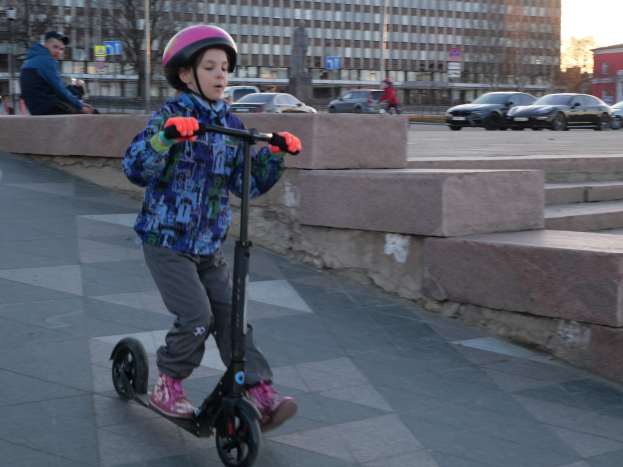 Ein junger Junge in einem Helm und Handschuhen fährt mit einem Roller eine Treppe auf einem Gehweg herunter, mit Fahrzeugen, Menschen, Bäumen, Polen, Brettern, Gebäuden und einem klaren blauen Himmel im Hintergrund.