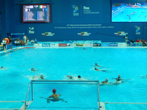 Wasserballspieler bei den 2016 FINA-Weltmeisterschaften in Kazan im Pool gegeneinander antretend, während links Zuschauer auf Stühlen sitzen und ein Scoreboard mit Event-Branding im Hintergrund angezeigt wird.