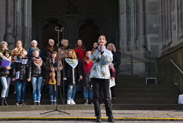 Eine Frau in der Mitte hält ein Mikrofon und singt, umgeben von einer Gruppe von Menschen mit Musikinstrumenten, vor einem Gebäude mit Stufen, Geländern und einer Wand mit Säulen und einer Tür.