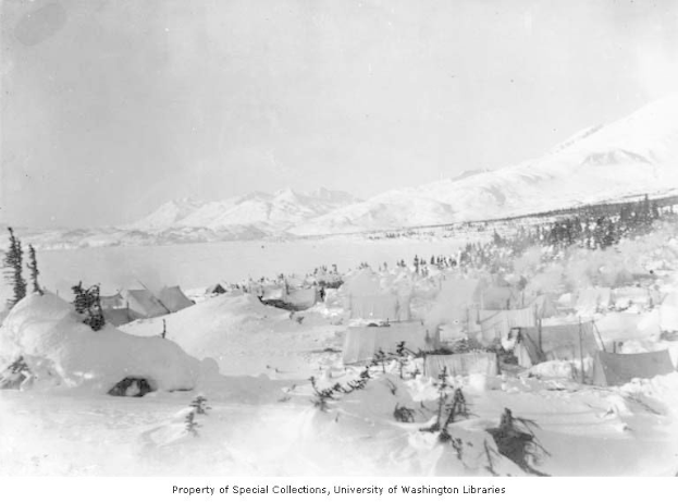 Schwarz-weiß-Foto einer Gruppe von Zelten im Schnee, umgeben von Bäumen und Bergen, mit dem Text "Kootenay Camp, Alaska, circa 1900" unten.