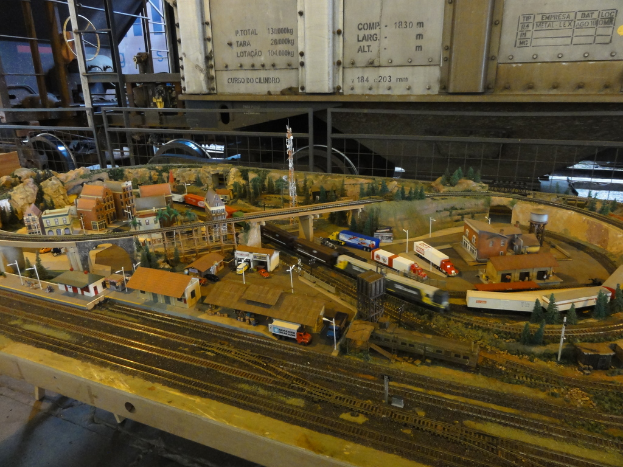 Eine Miniaturszene auf einem Tisch mit einer Bahnschiene, einem Zug, Gebäuden, Fahrzeugen, einer Brücke, einem Turm und Bäumen.