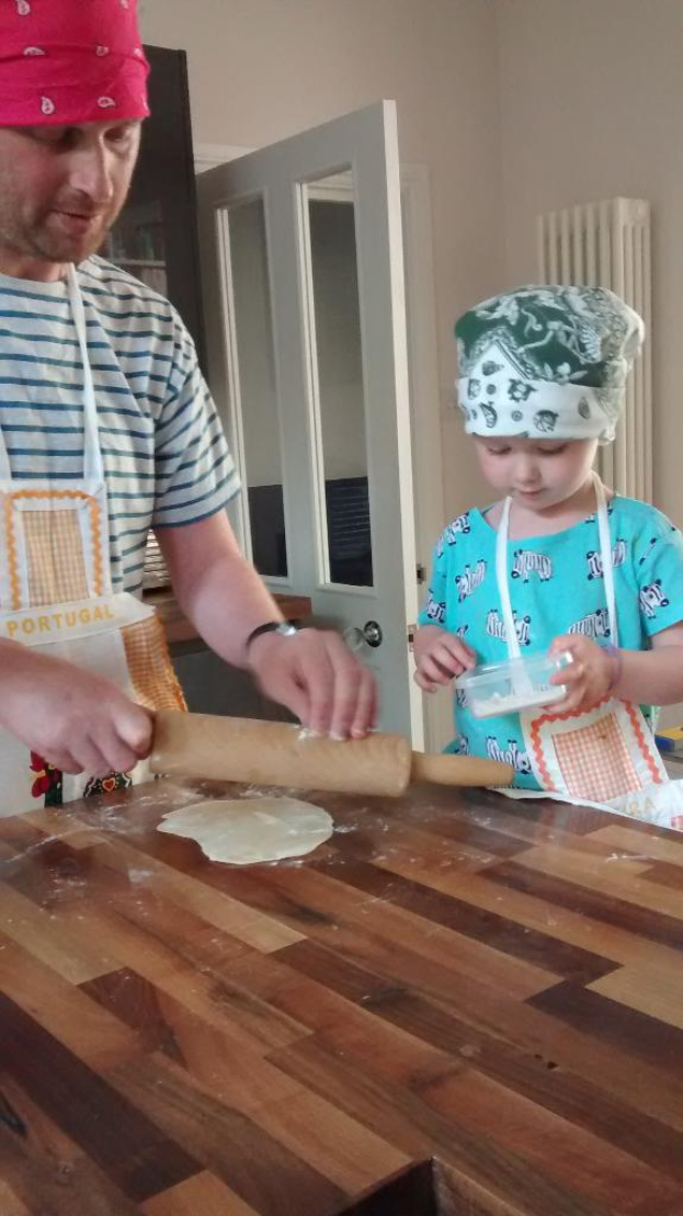 Ein Mann und ein Kind, beide mit Schürzen und Mützen bekleidet, stehen vor einer Tür mit Griff; der Mann hält eine Rolle, während das Kind einen Behälter hält und ein Tisch mit Essen zwischen ihnen sichtbar ist.
