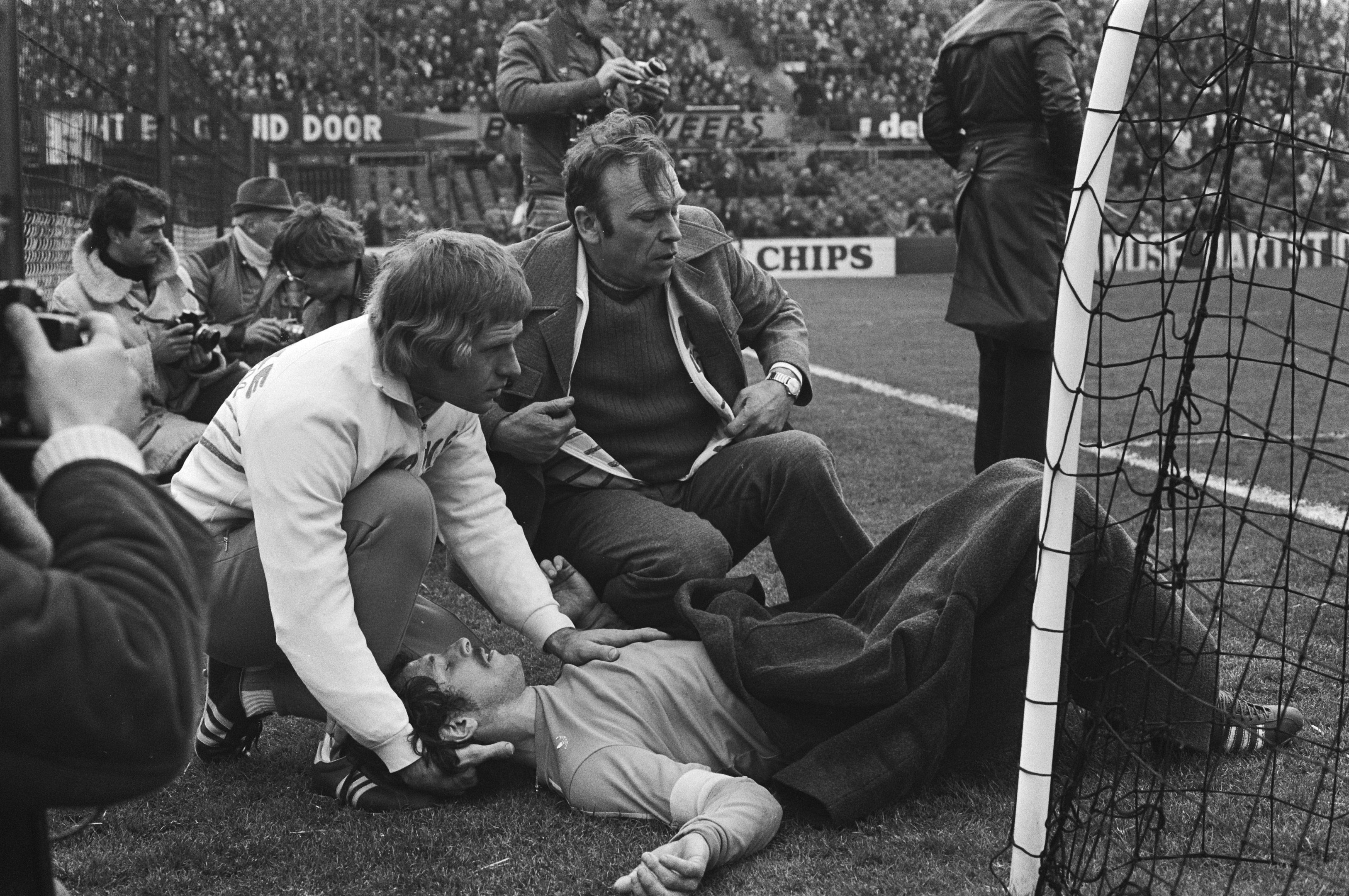 Ein Mann liegt neben einem Fußballtor auf dem Boden, umgeben von sitzenden und stehenden Zuschauern auf dem Rasen, mit einem Netz auf der rechten Seite und einer Menge Zuschauern in den Tribünen dahinter.