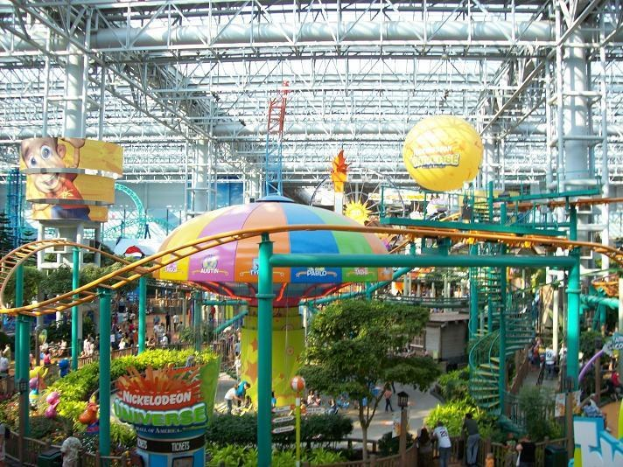 Großes Indoor-Wasserpark mit einer Achterbahn, die durch die Mitte verläuft und von Menschen auf Wasserattraktionen umgeben ist, umgeben von Grünflächen und beleuchtet von Deckenlampen und Stangen.