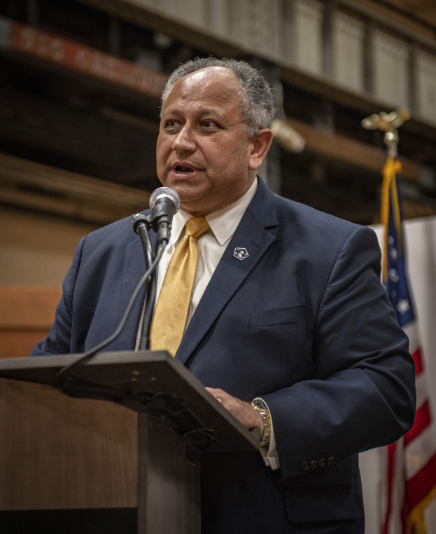 Ein Mann in Anzug und Krawatte spricht an einem Podium mit Mikrofon, rechts ein Fahne und eine Wand im Hintergrund, wahrscheinlich spricht er zur Ankündigung des neuen Bürgermeisters von New York City.