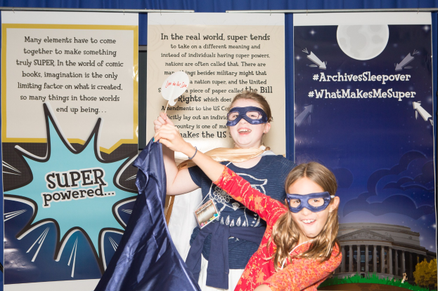 Zwei junge Mädchen in Superheldenkostümen posieren für ein Foto vor einem blauen Vorhang, eine hält ein Tuch, während beide Masken tragen, mit Bannern im Hintergrund, die auf ein Schulfest hinweisen.