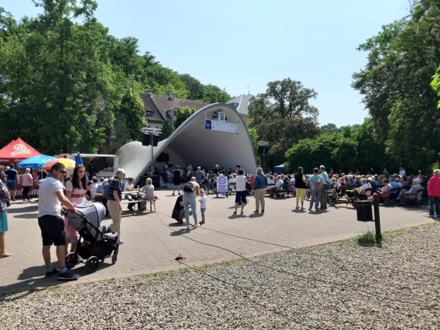 Eine Menschenmenge versammelt sich vor einem Gebäude bei der Festival-Eröffnung, einige sitzen auf Bänken und andere stehen; ein Kinderwagen ist im Vordergrund zu sehen, sowie Bäume, Gebäude und ein klarer blauer Himmel im Hintergrund. Zelte säumen die linke Seite, und ein Mülleimer ist auf der rechten Seite zu sehen.