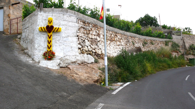 Eine Straße auf der rechten Seite mit Pflanzen, einem Pfahl, einer Fahne, einem christlichen Schild und Blumen; eine Steinmauer, Bäume, Drähte, ein Tor auf der linken Seite und Gebäude im Hintergrund.