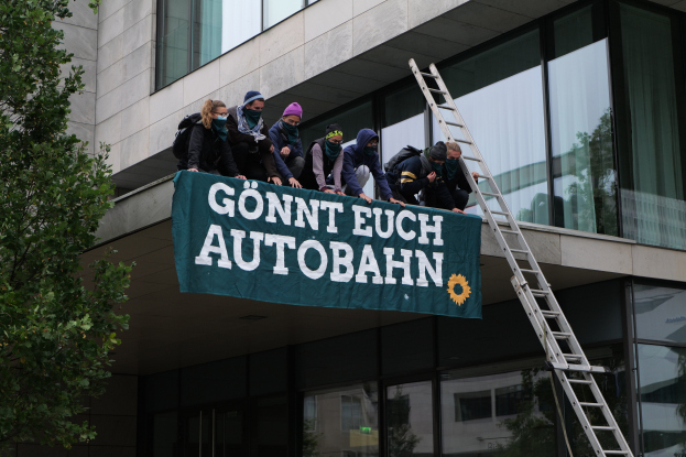 Eine Gruppe von Menschen auf dem Dach eines Gebäudes stehend, eine Fahne haltend, mit einem Baum links und Glasfenstern im Hintergrund, wahrscheinlich im Support der deutschen Regierung protestierend.