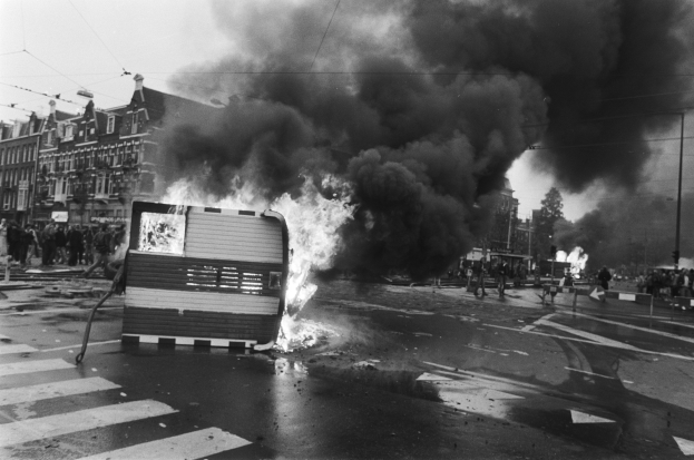 Schwarzes und weißes Foto eines brennenden Busses am Straßenrand mit Menschen in der Nähe, umgeben von Pfosten, einem Schild, Bäumen, Gebäuden und einem bewölkten Himmel.