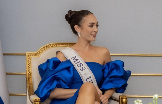 Frau in einem blauen Kleid mit einem "Miss Universe 2019"-Band, die auf einem Stuhl sitzt und lächelt, umgeben von Blumen.