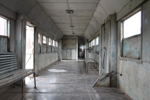 Innensicht eines alten Zugwagens mit Holzsitzen, Fenstern auf beiden Seiten und einer Decke, mit einem Gebäude im Hintergrund.