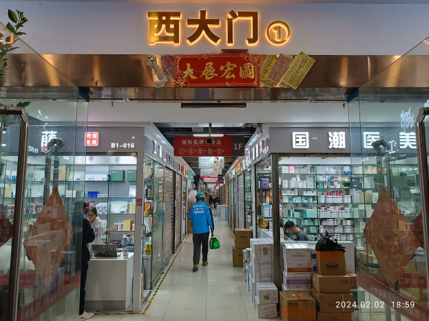 Innenansicht einer chinesischen Apotheke mit Glaswänden, Regalen voller Artikel, verstreuten Kartons, einer Pflanze links und einer Namensplakette oben.