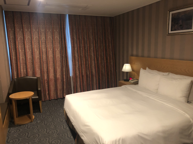 Hotelzimmer mit großem Bett, Stuhl, Tisch, Telefon, Lampe, Foto auf dem Wand, Fenster mit Vorhängen und Deckenbeleuchtung.