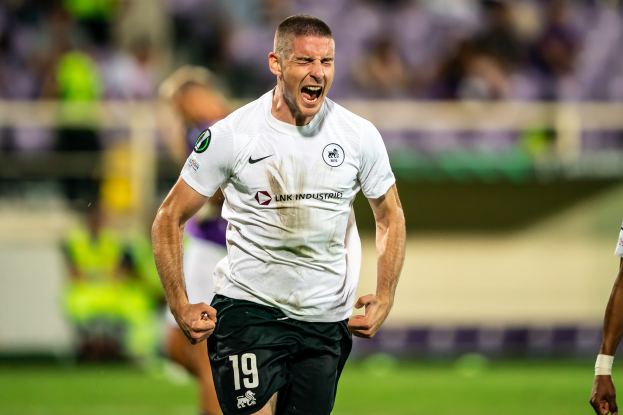 Ein Mann in weißem Hemd und schwarzen Shorts feiert auf einem Fußballfeld, umgeben von einer Gruppe von Menschen, wahrscheinlich nach einem Tor in einem Spiel zwischen Leeds United und Melbourne Victory.