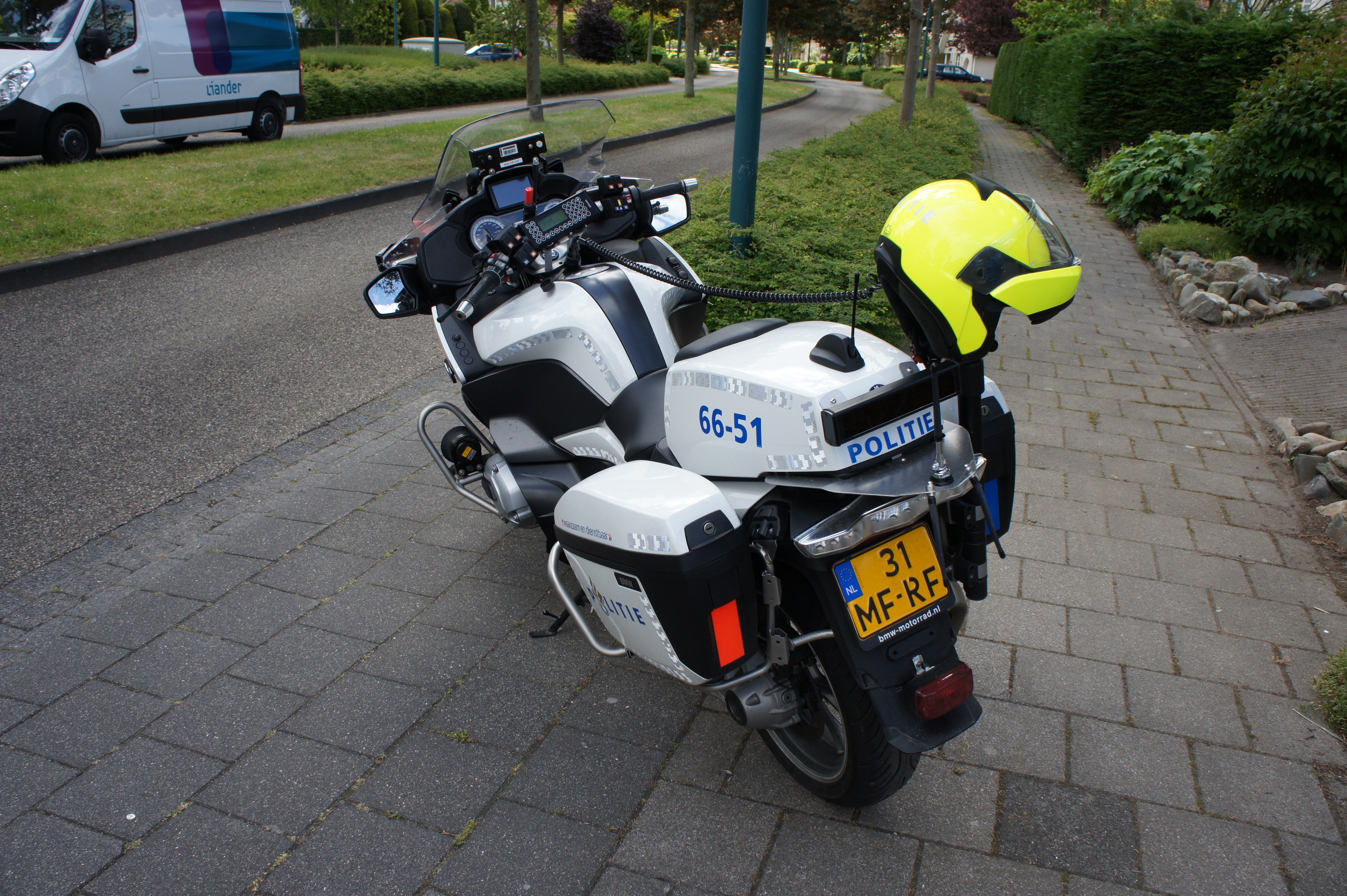 Ein polizeiliches Motorrad am Straßenrand mit einem Helm darauf, umgeben von Bäumen, Masten, Gebäuden, Pflanzen, Gras, Steinen und anderen Fahrzeugen.