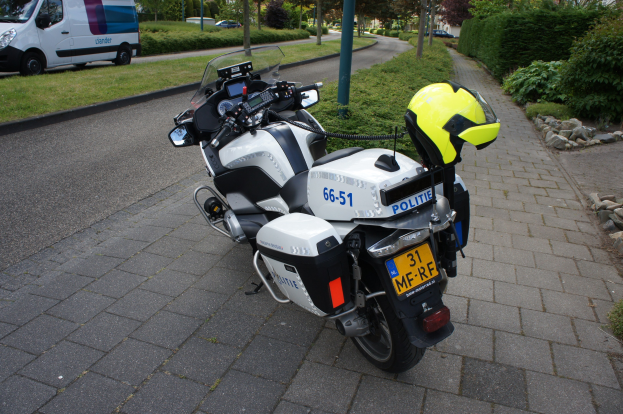 Ein polizeiliches Motorrad am Straßenrand mit einem Helm darauf, umgeben von Bäumen, Masten, Gebäuden, Pflanzen, Gras, Steinen und anderen Fahrzeugen.
