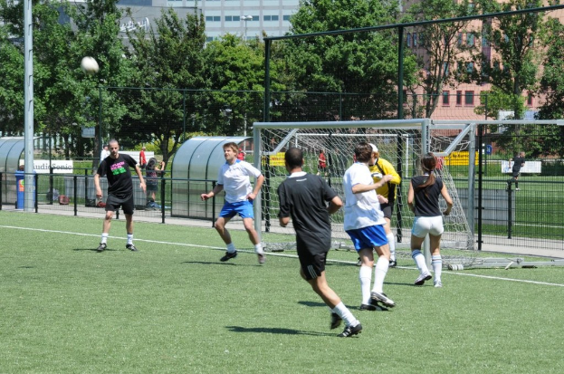 Personen in Kleidung beim Fussballspielen auf einem Feld mit einem Zaun und einem Tor in der Mitte, einem Pfosten und einem Fussball oben links und Bäumen und Gebäuden im Hintergrund.