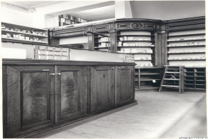 Ein Schwarz-Weiß-Foto eines Apothekeninneren mit einem Tresen, Schränken, einer Leiter und Regalen, die mit verschiedenen Gegenständen gefüllt sind, mit Text oben auf dem Bild.
