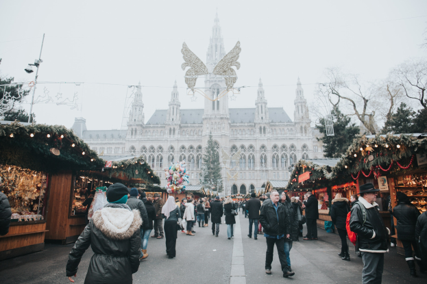 Ein lebendiger Weihnachtsmarkt in Wien, Österreich, mit Menschen, die herumlaufen, Ständen, die mit Lichtern und festlichen Décorations geschmückt sind, einem Gebäude mit Fenstern im Hintergrund, Bäumen und einem klaren blauen Himmel.