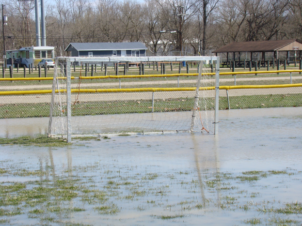 Ein Fußballtor steht im Zentrum eines überfluteten Feldes mit Gras und Wasser drumherum, vor dem Hintergrund von Hütten, Pfählen, Bäumen, Fahrzeugen und einem klaren blauen Himmel.
