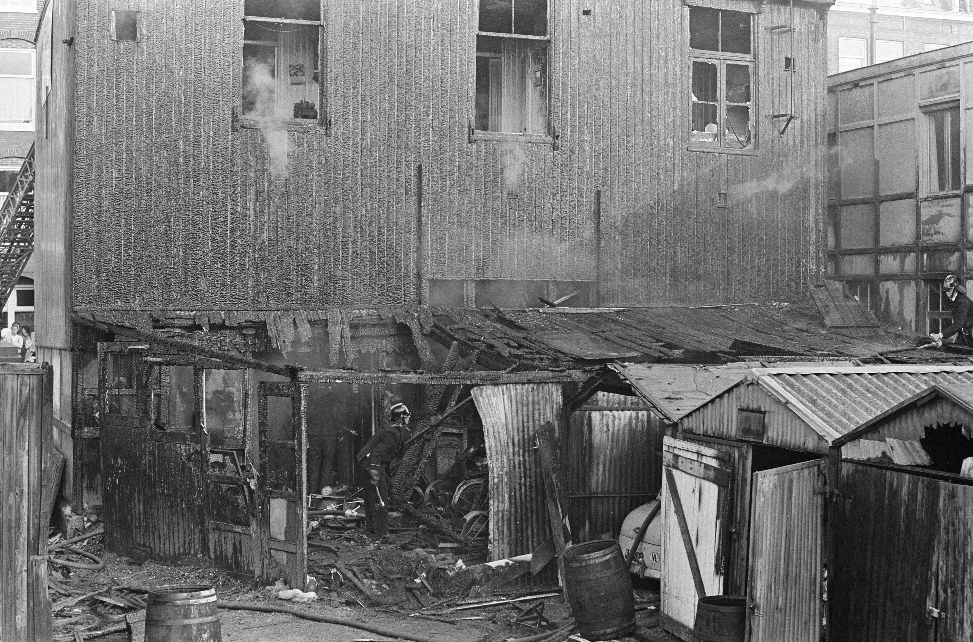 Schwarzes und weißes Foto eines feuerbeschädigten Gebäudes mit verstreuten Trümmern, darunter Fässer und hölzerne Gegenstände, sowie einigen Menschen in der Nähe, mit anderen Gebäuden, einer Leiter und einem Pfahl im Hintergrund.
