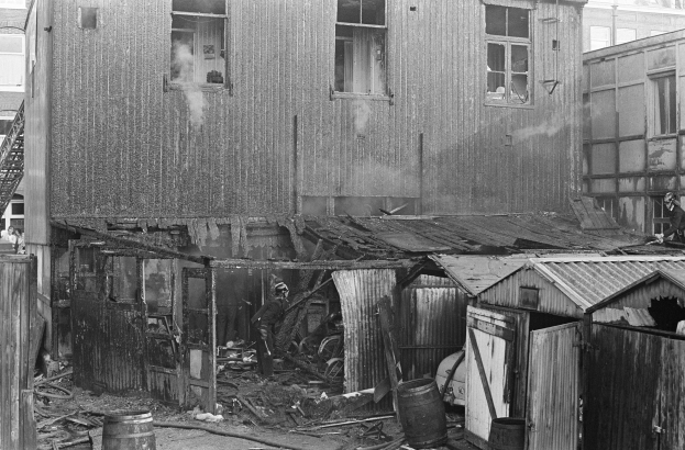 Schwarzes und weißes Foto eines feuerbeschädigten Gebäudes mit verstreuten Trümmern, darunter Fässer und hölzerne Gegenstände, sowie einigen Menschen in der Nähe, mit anderen Gebäuden, einer Leiter und einem Pfahl im Hintergrund.