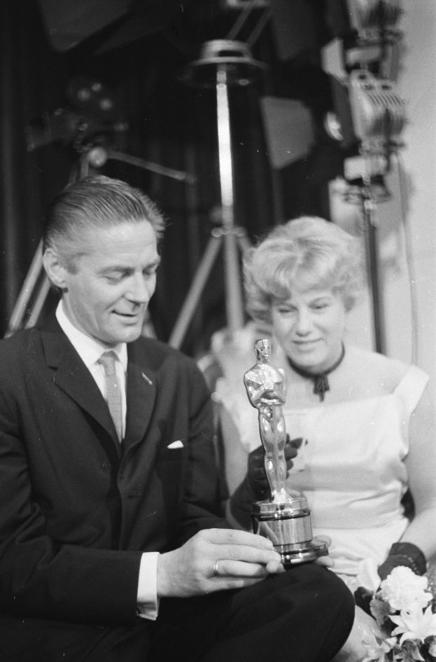 Ein Mann in einem schwarzen Blazer, weißem Hemd und Krawatte hält eine Oscar-Statue, während er neben einer Frau in Abendgarderobe sitzt, die einen Blumenstrauß hält, wobei das Schwarz-Weiß-Szenario von Lichtern beleuchtet wird.