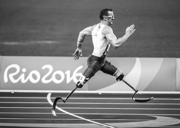 Ein Mann mit einer Prothese rennt auf einer Laufbahn, im Hintergrund ist ein Schild zu sehen mit der Aufschrift "Rio 2016 Paralympische Spiele", dargestellt in Schwarz-Weiß.