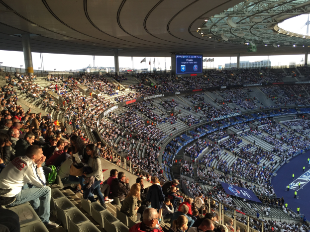 Große Menschenmenge in einem Stadion bei einem Fußballspiel mit einer Bühne auf der rechten Seite, Fahnen, Stangen, einem Bildschirm und der Allianz Arena in München, Deutschland im Hintergrund.