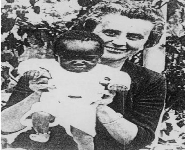 Ein Schwarz-Weiß-Foto einer Frau, die strahlend ihr Baby hält, mit Blättern im Hintergrund.