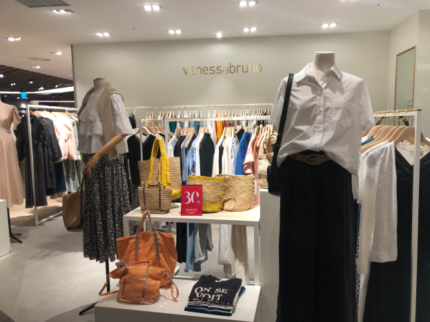 Ein Vanessa Bruno-Laden in Singapur mit Schaufensterpuppen in Kleidern, Kleidung an Bügeln, Taschen und anderen Gegenständen auf Tischen, einer Texttafel an der Wand, einem Spiegel auf der rechten Seite und Deckenleuchten.