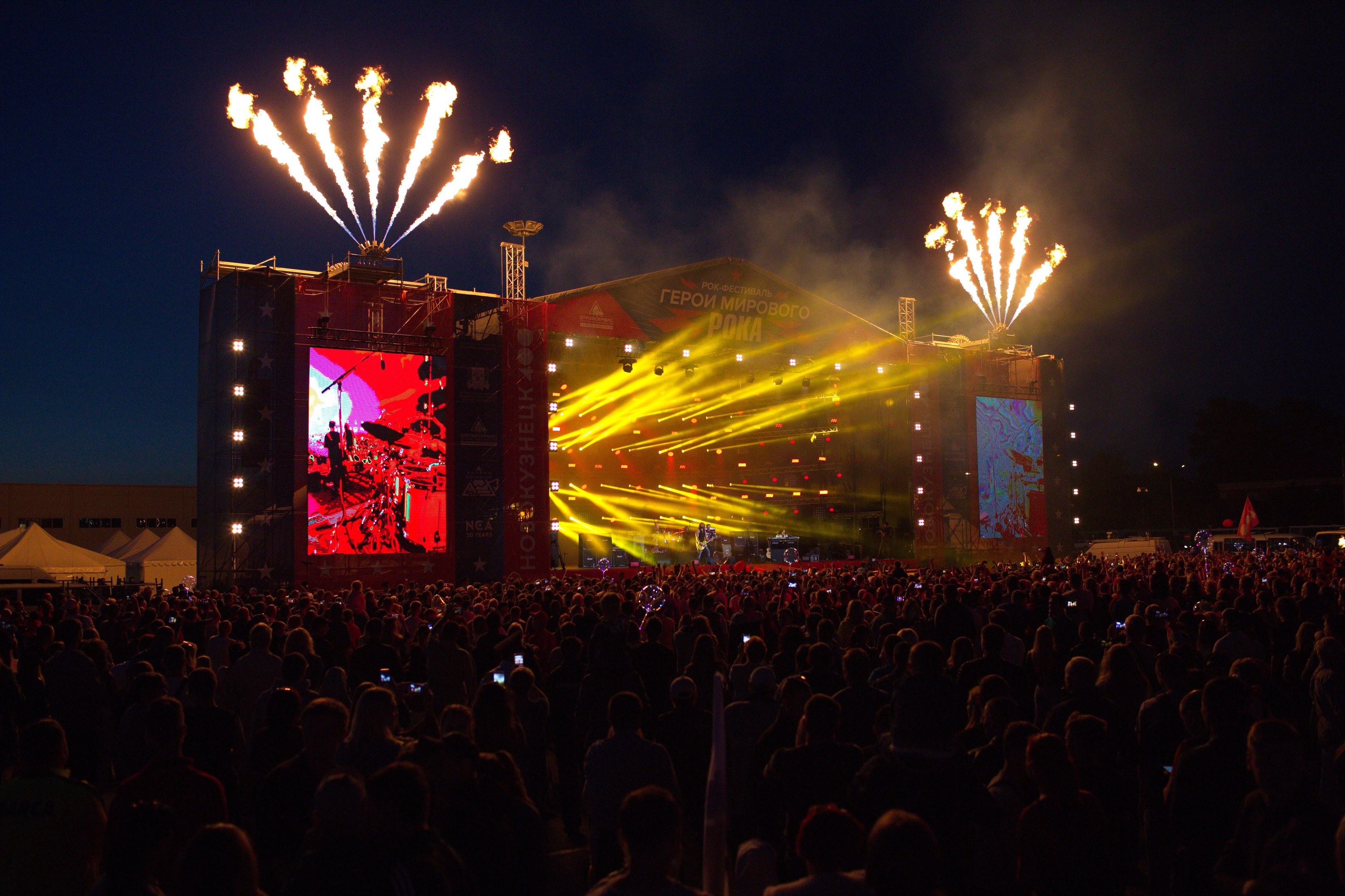 Eine große Menschenmenge auf einem Musikfestival schaut auf einer hell erleuchteten Bühne performende Künstler, mit Zelten links und Feuerwerk im dunklen Himmel dahinter.